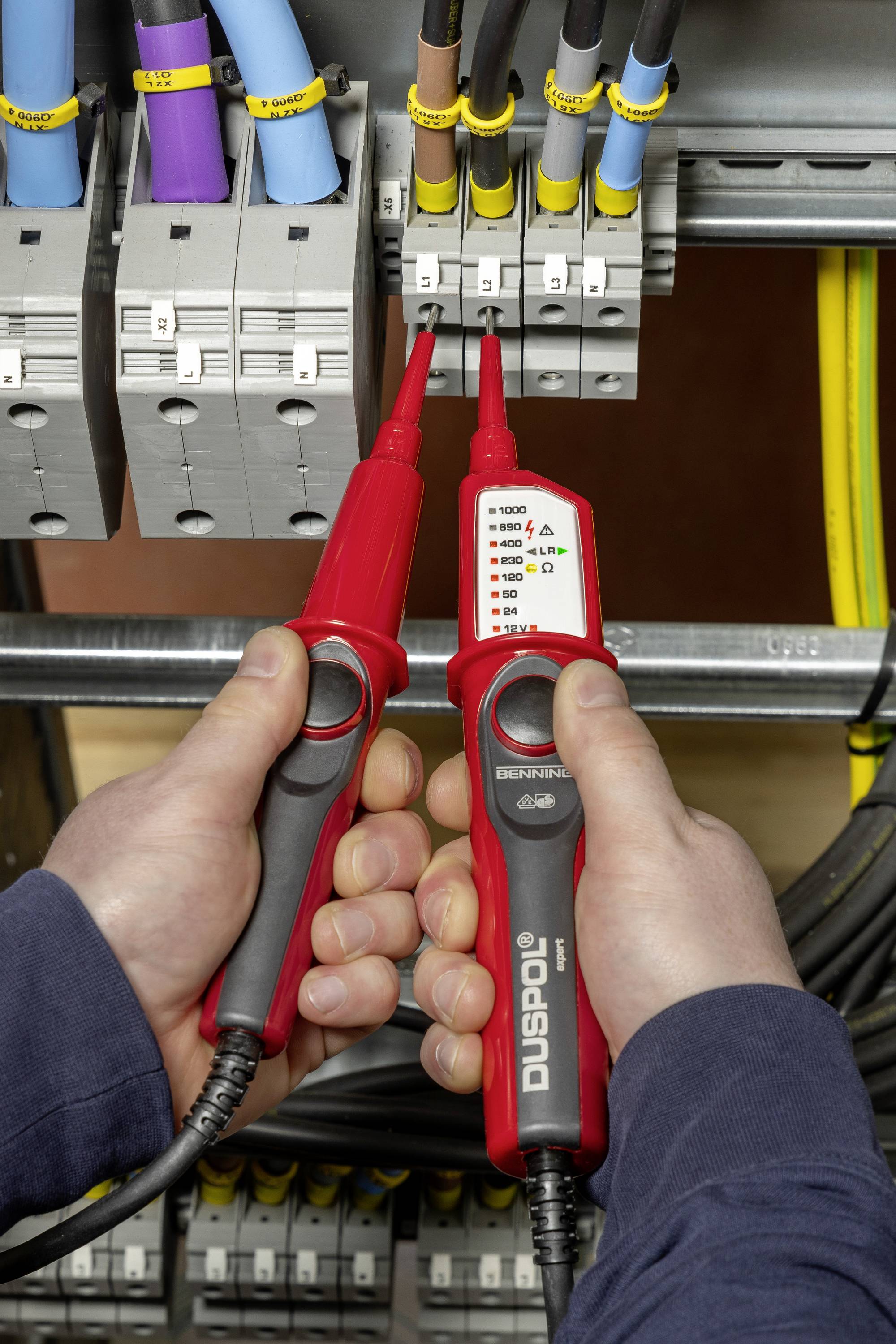 Dvě ruce drží měřicí přístroj napětí, aby zkontrolovaly elektrický proud v rozvaděči. Je připojeno několik kabelů.