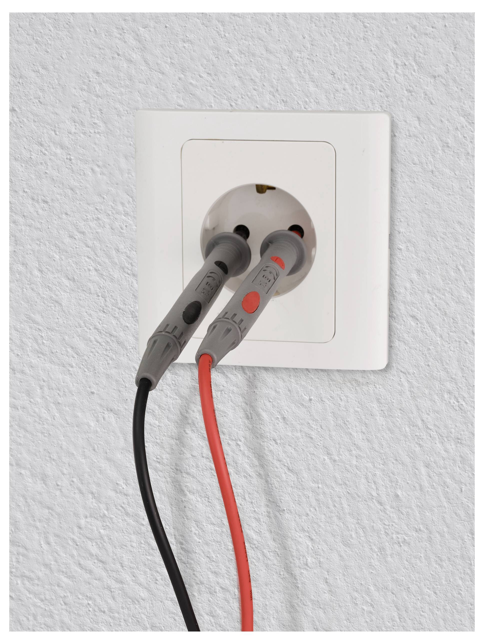 Elektrická zásuvka se dvěma vloženými sondy, jednou červenou a jednou černou, pravděpodobně testující napětí. Bílé pozadí stěny.