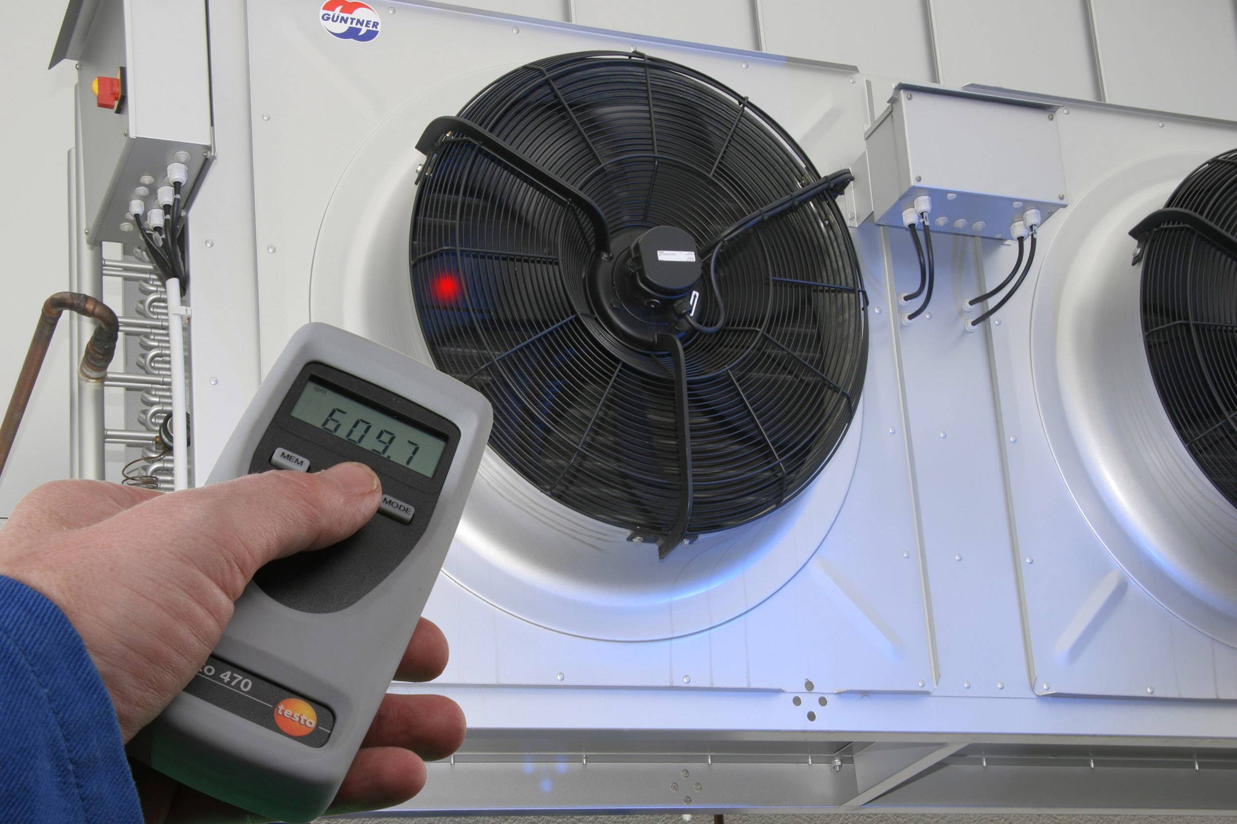 Ruční přístroj měří otáčky velkého průmyslového ventilátoru na vnější stěně. Jsou viditelná dvě velká tělesa ventilátorů.