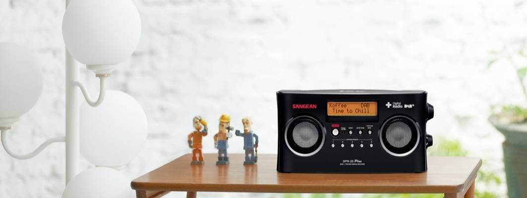 Černé rádio stojí na dřevěném stole vedle tří figurek. Na displeji svítí nápis 'Time to Chill'. Vlevo v pozadí jsou vidět lampy.