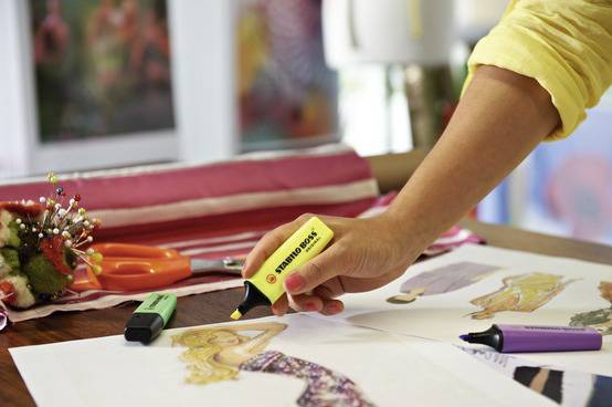 Osoba zvýrazňuje módní nákresy žlutým textikovým zvýrazňovačem. V pozadí jsou vidět barevné látky a špendlíky.