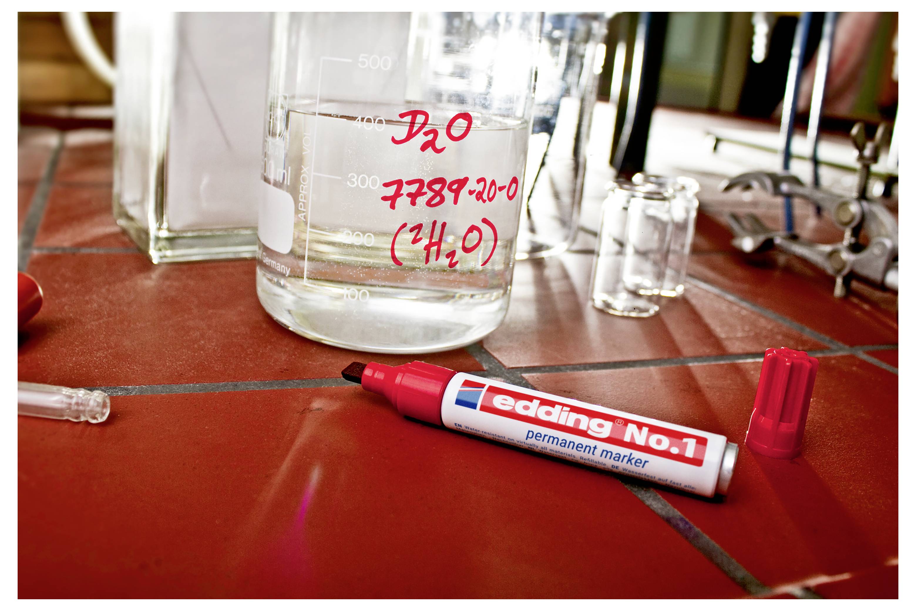 Červený permanentní fix leží na dlaždičkovém laboratorním stole vedle skleničky označené "D2O 7789-20-0 (H2O)".