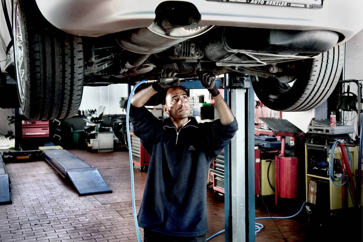 Mechanik pracuje pod zvednutým autem v autoservisu, obklopen nástroji a zvedáky.