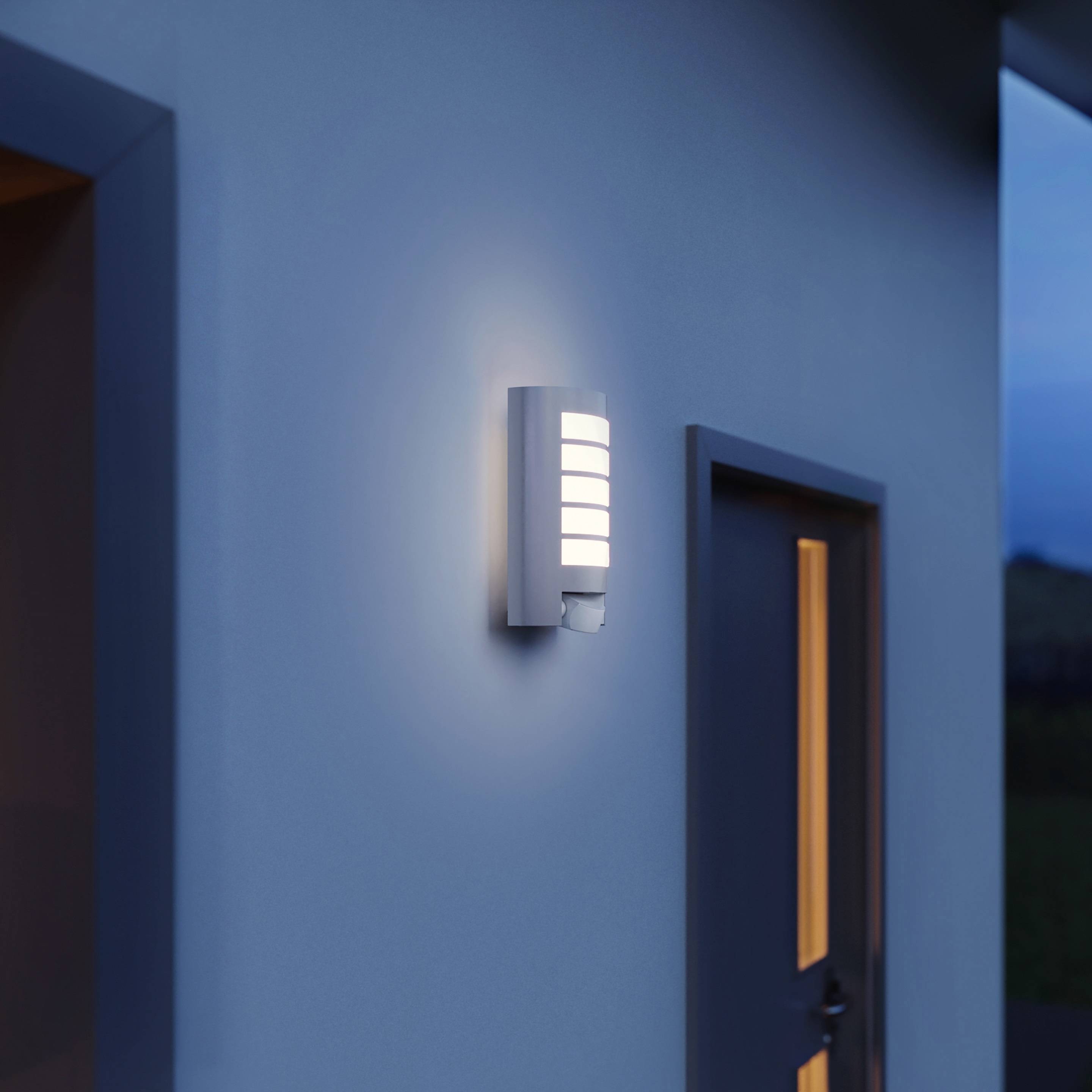 Vnější stěna moderní nemovitosti s rozsvíceným nástěnným svítidlem vedle dveří; atmosféra soumraku.