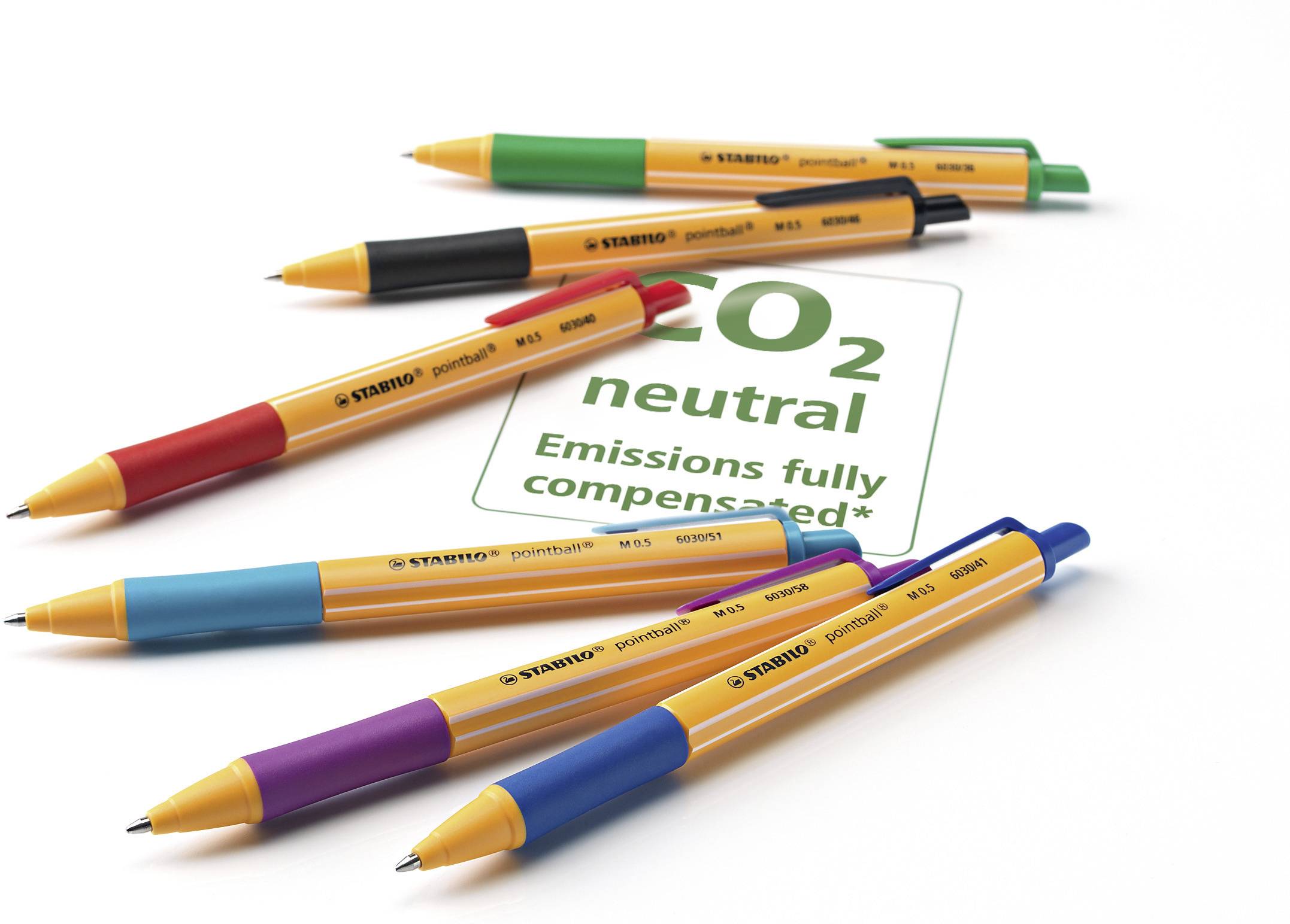 Šest barevných per obklopuje ceduli s nápisem 'CO2 neutrální, emise plně kompenzovány', na bílém pozadí.