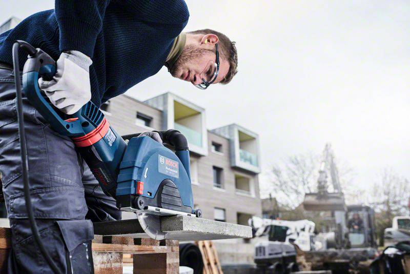 Dělník používá elektrickou úhlovou brusku k rozřezání kovového nosníku na staveništi. V pozadí jsou viditelné moderní budovy.