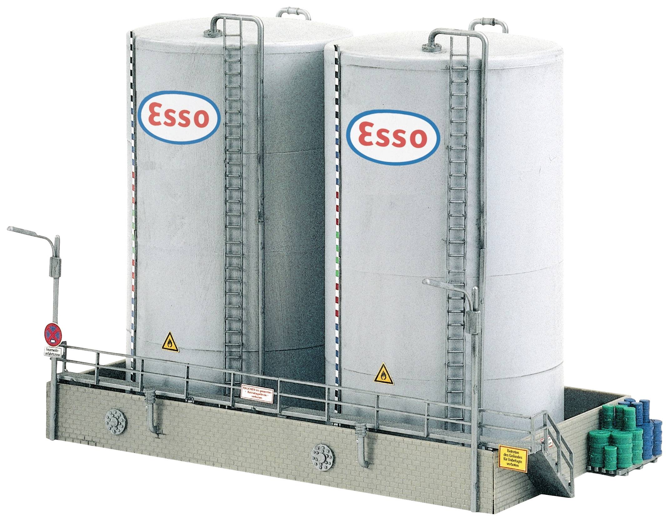 Dva velké bílé tanky s logem 'Esso', propojené žebříky a potrubím, stojí na plošině. Vedle tanků jsou modré a zelené sudy.
