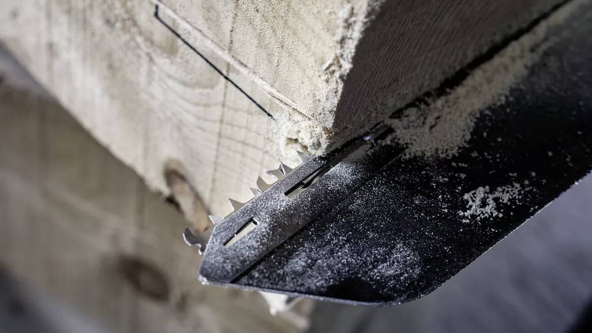 Detailní záběr ruční pily, která řeže hrubou dřevěnou deskou. Viditelné piliny ukazují ruční opracování dřeva.