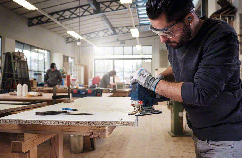 Muž v ochranných brýlích a rukavicích řeže dřevo přímočarou pilou v dílně. V pozadí pracují další osoby.