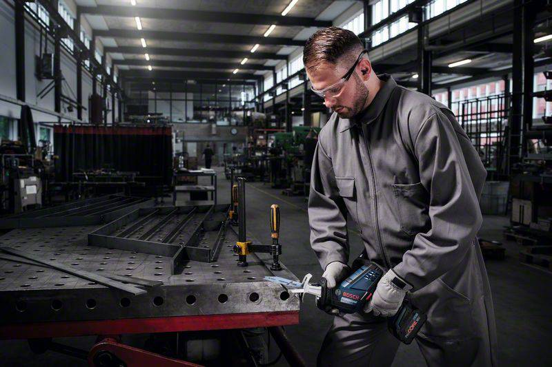 Dělník v šedém ochranném obleku a ochranných brýlích pracuje s elektrickým nástrojem u velkého kovového stolu v dílně.