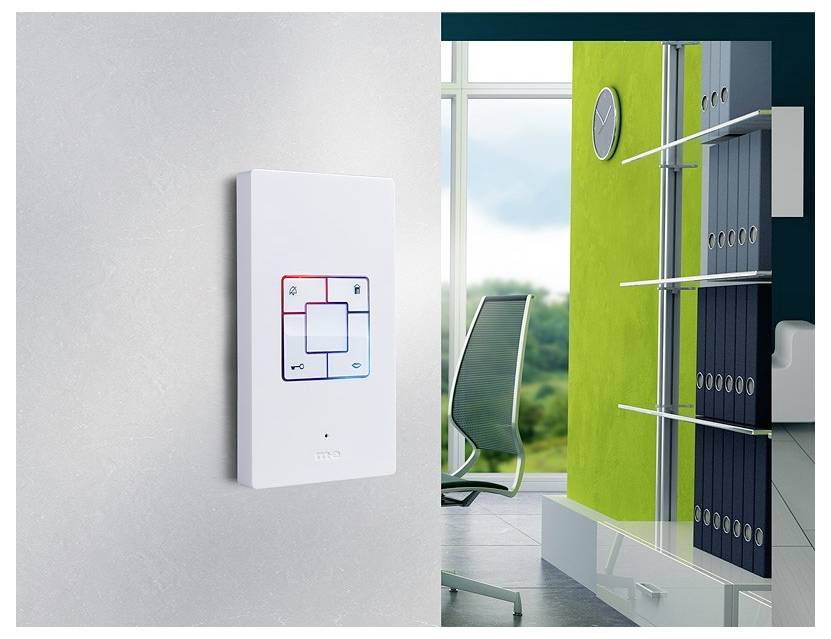 Osvětlený elektronický nástěnný vypínač s červeným a modrým světlem; moderní kancelářská oblast v pozadí se židlí a zelenou stěnou.