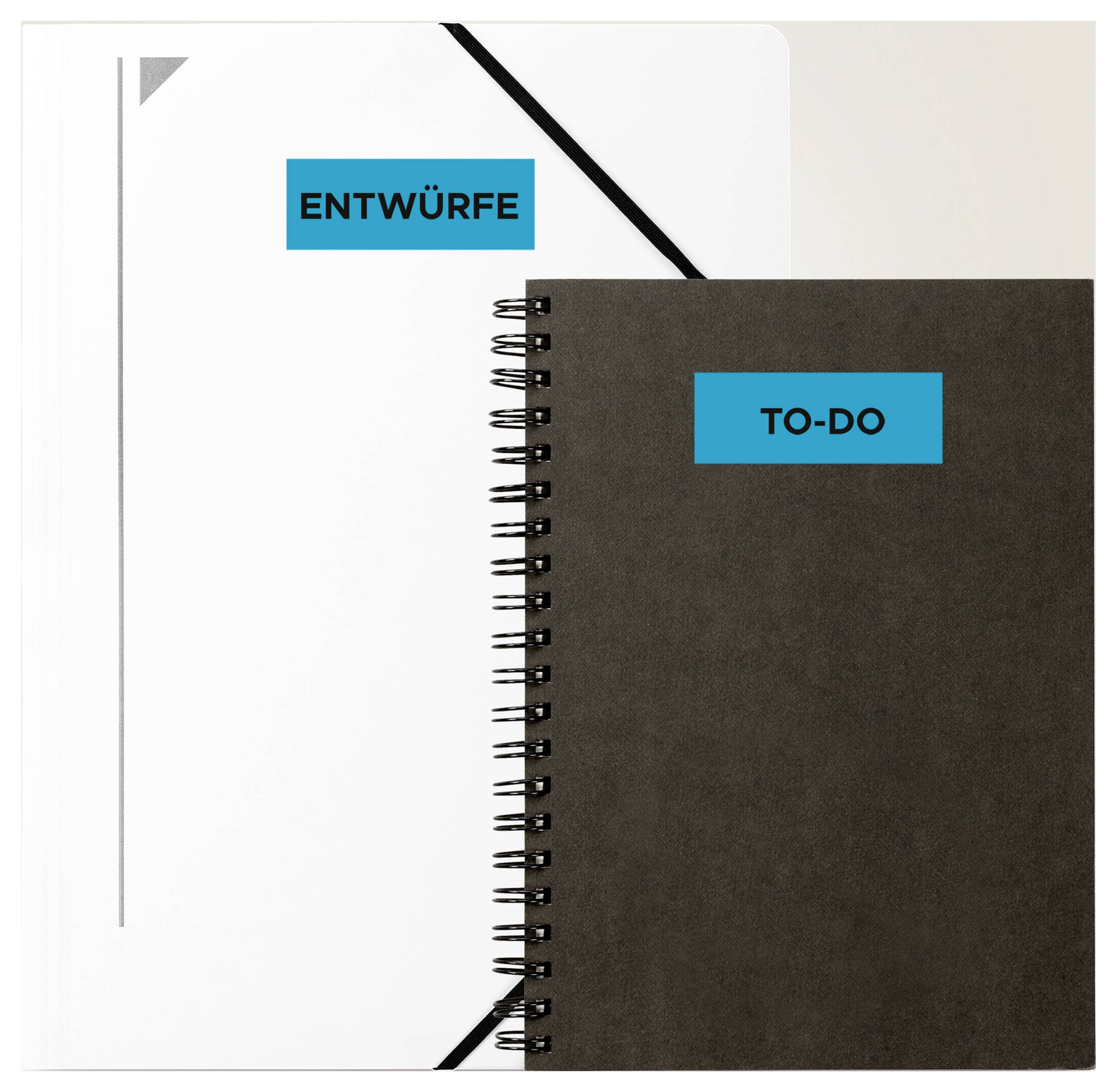Dva spirálem vázané notebooky, vlevo bílý s nálepkou "NÁVRHY", vpravo tmavý s nálepkou "ÚKOLY", uspořádané vedle sebe s částečným přesahem.