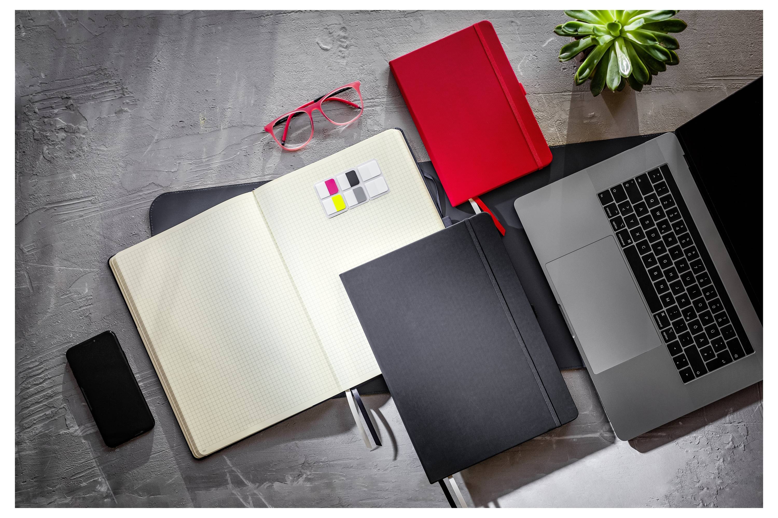 Pohled shora na kancelářský stůl s otevřeným notebookem, laptopem, složkami, smartphonem, červenými brýlemi, lepicími poznámkami a malou rostlinou.