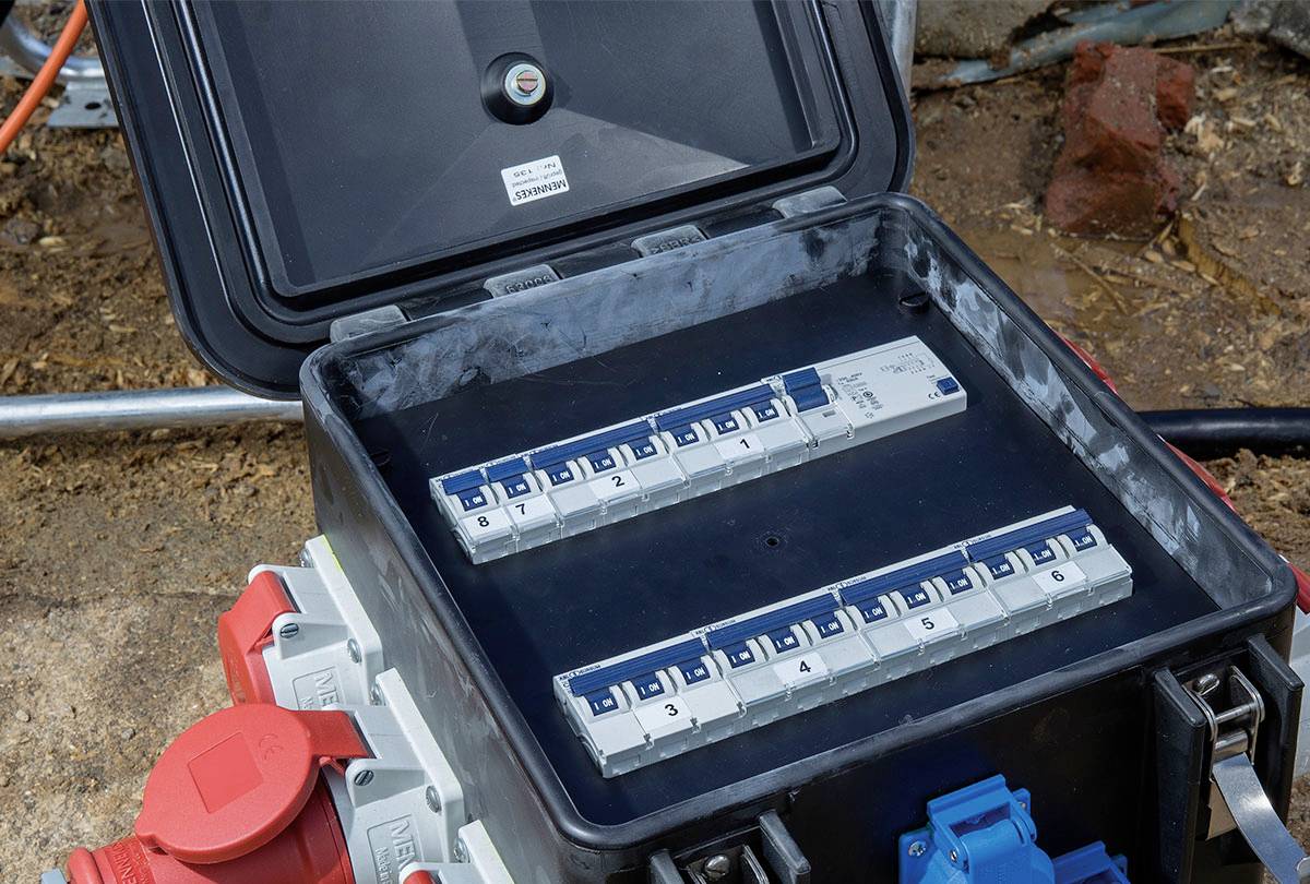 Otevřená elektrická rozvodná skříň s několika modrými jističi s čísly. Tři červené a jedna modrá zásuvka po straně.