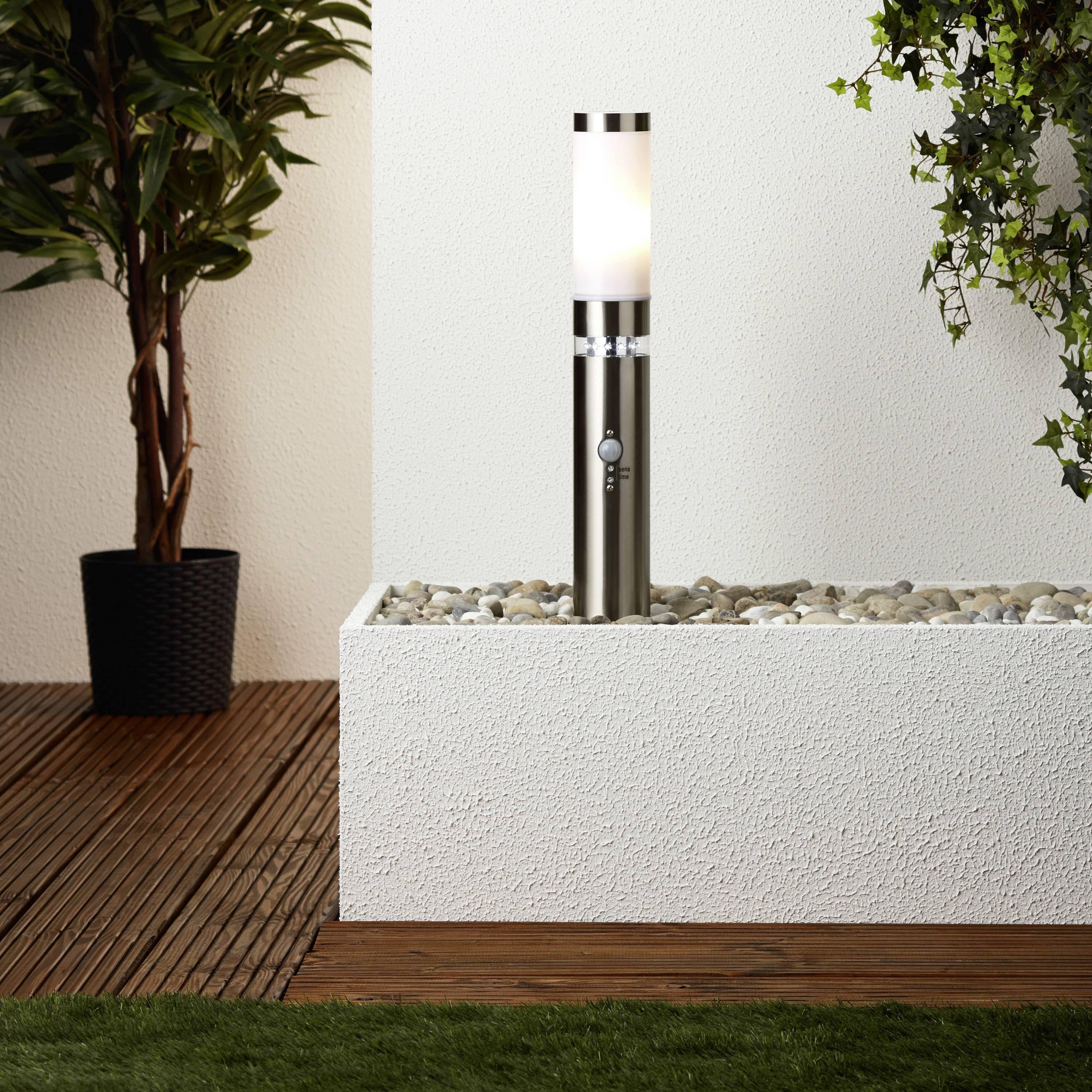 Moderní stojací lampa stojí na nízkém podstavci z oblázků před bílou stěnou, v pozadí je umístěn květináč s rostlinou.