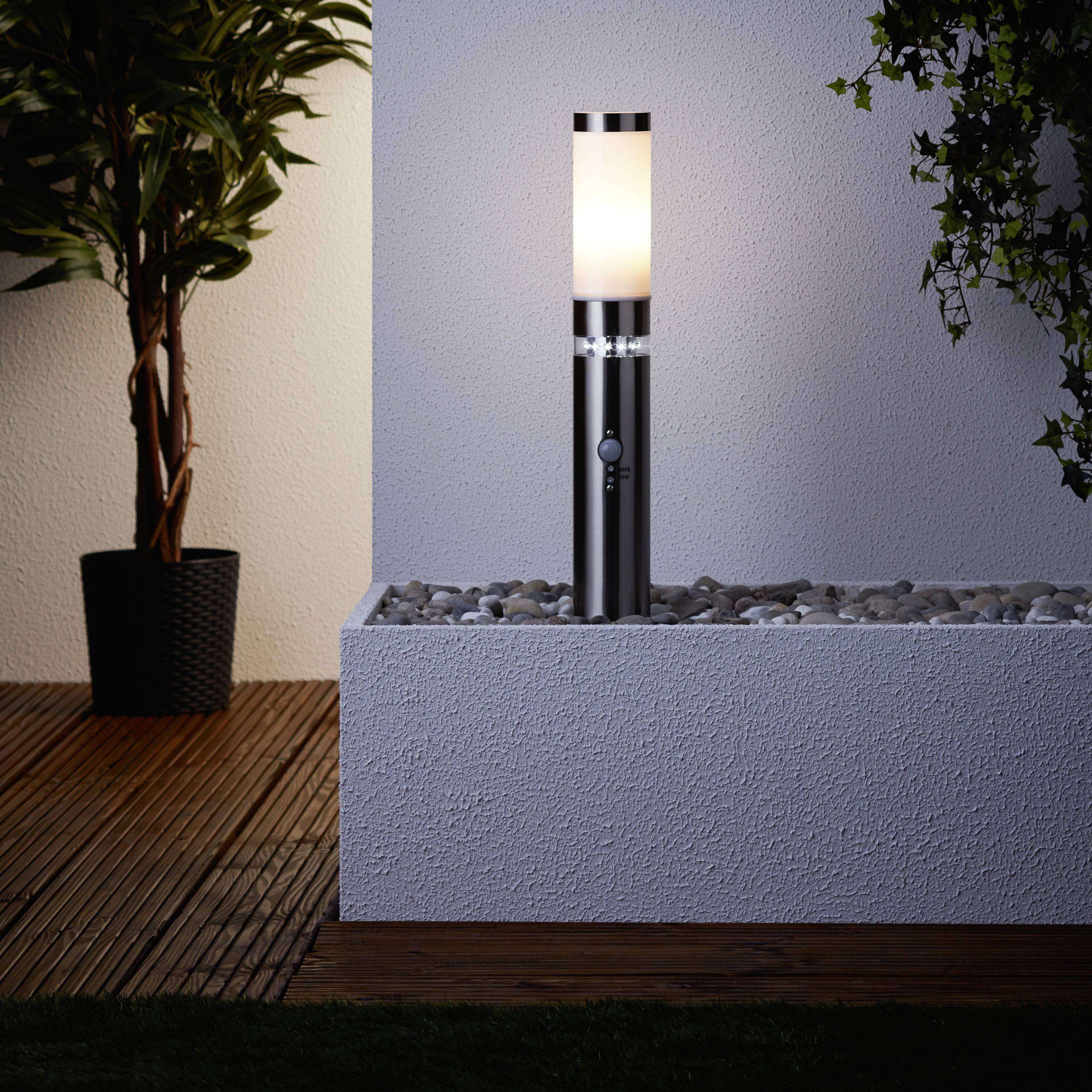 Moderní stojací lampa ve venkovním prostoru stojí na podstavci, obklopena oblázky a rostlinami, osvětluje dřevěnou podlahu.