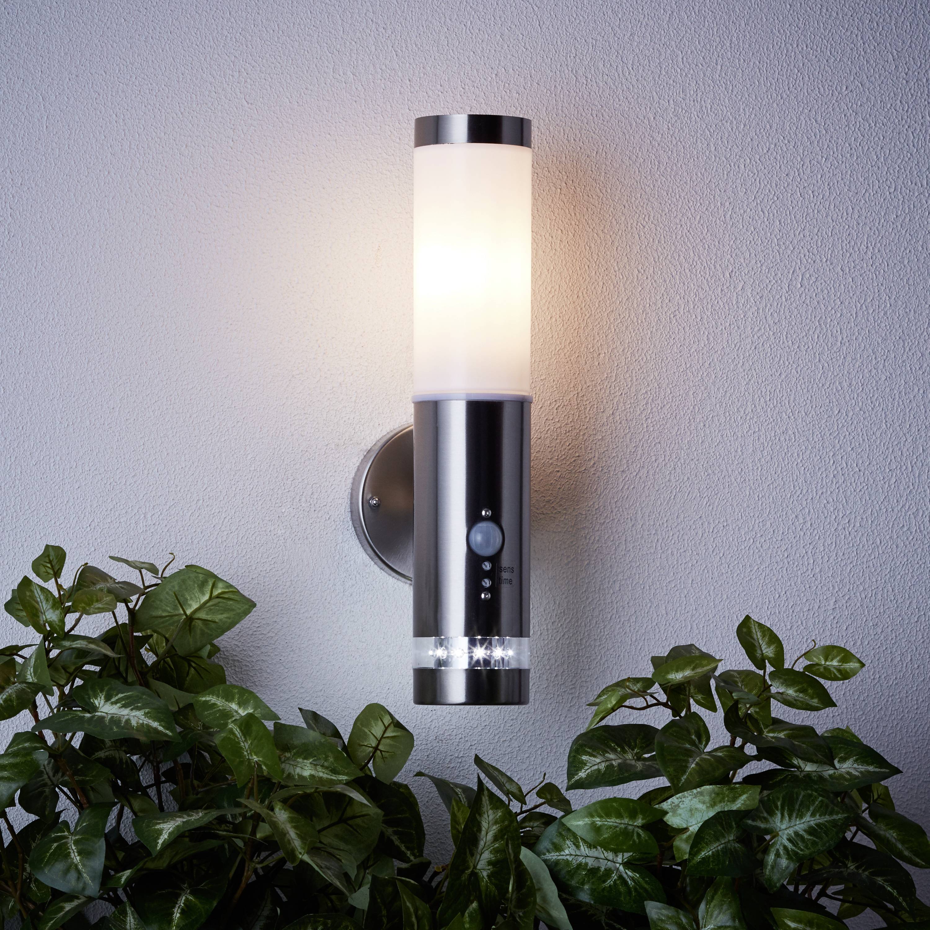 Nástěnná lampa v moderním designu svítí na bílé stěně, obklopená zelenými rostlinami.