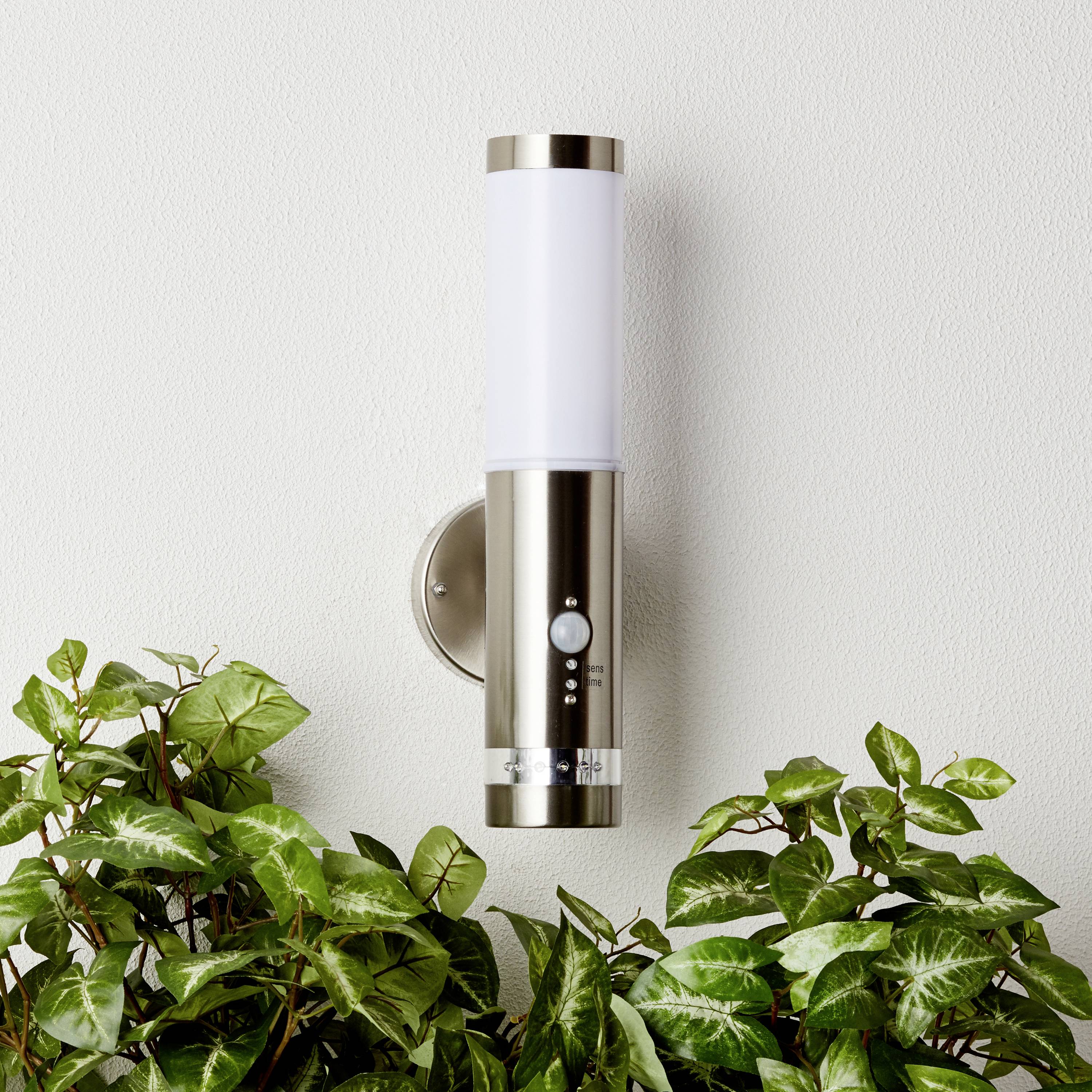 Moderní nástěnná lampa v designu nerezové oceli, upevněná na bílé stěně, obklopená zelenými rostlinami.