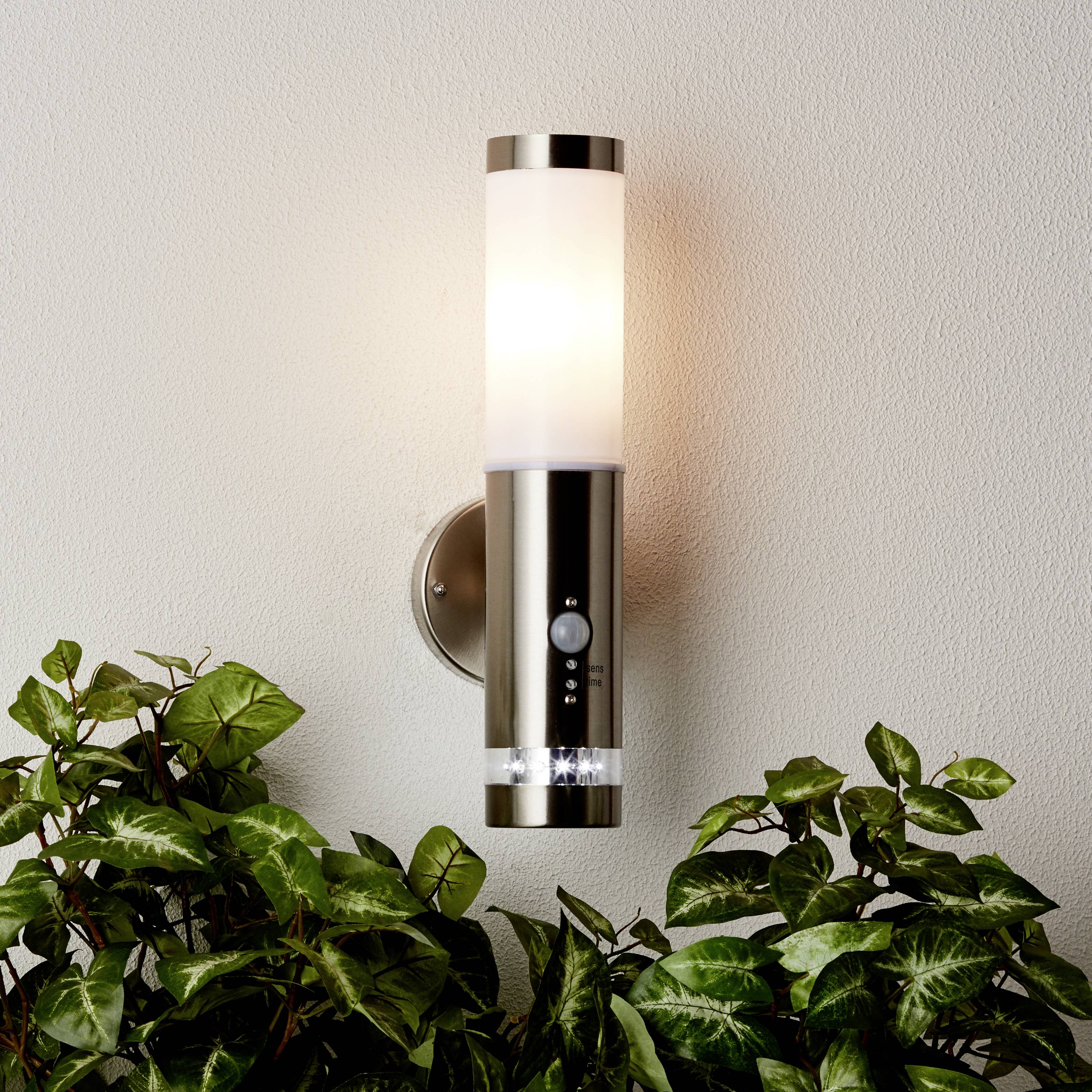 Moderní nástěnná lampa s válcovitým, lesklým tělem osvětluje prostor, umístěná na světlé stěně nad zelenými rostlinami.