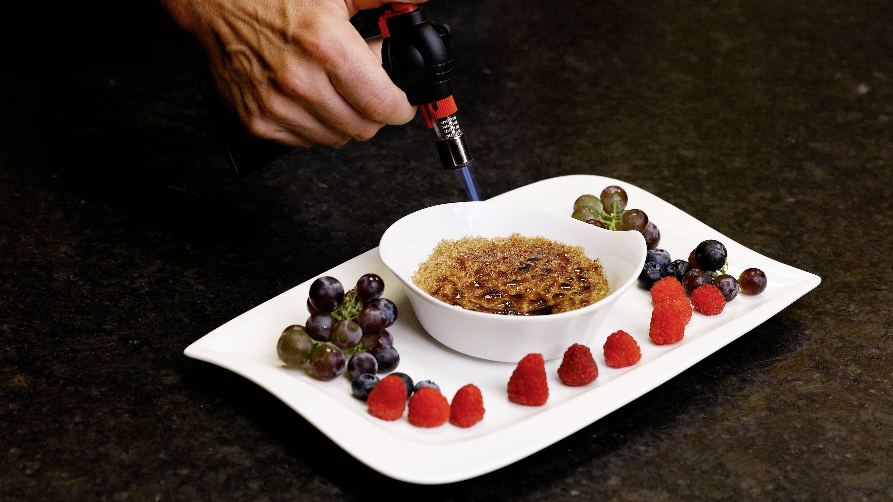 Ruka karamelizuje cukrovou krustu na crème brûlée pomocí kahanu. Talíř je ozdoben čerstvým hroznových a malinami.