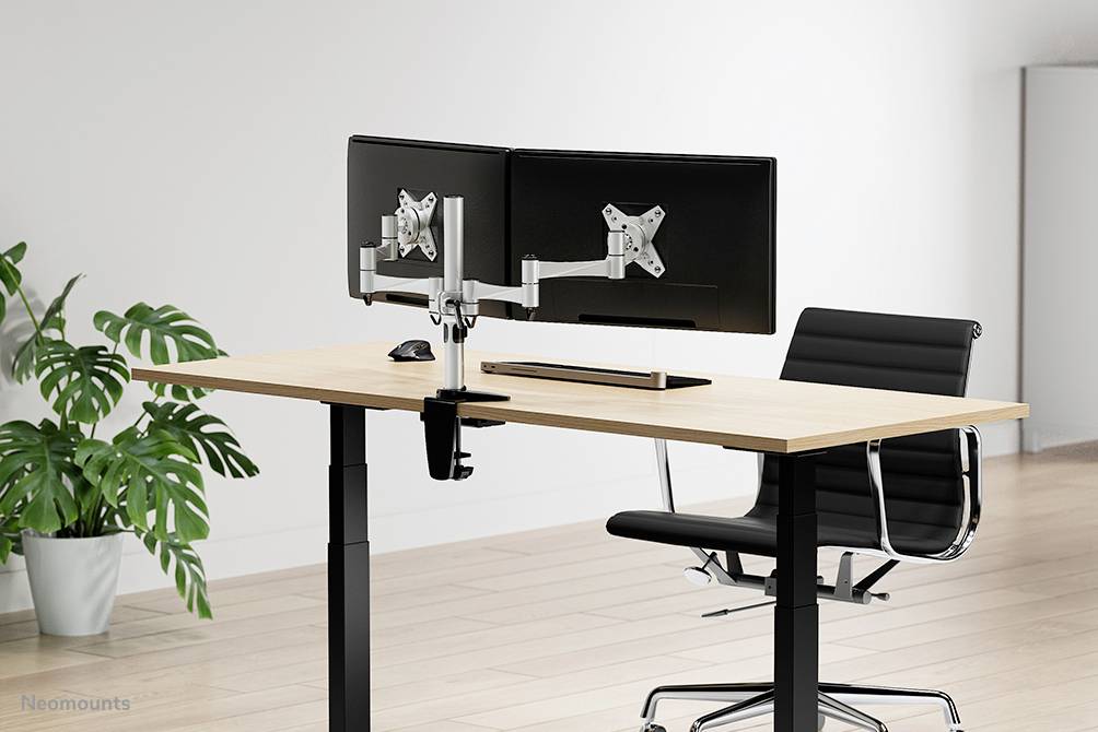 Moderní psací stůl se dvěma monitory, nastavitelným držákem a ergonomickou židlí na světlé dřevěné podlaze. Rostlina v pozadí.