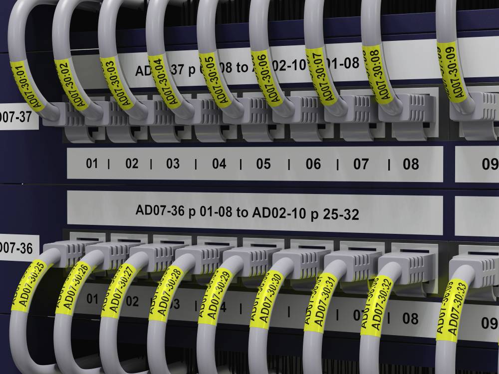 Serverová skříň s několika propojenými ethernetovými kabely, které jsou označeny žlutými štítky s alfanumerickými kódy.