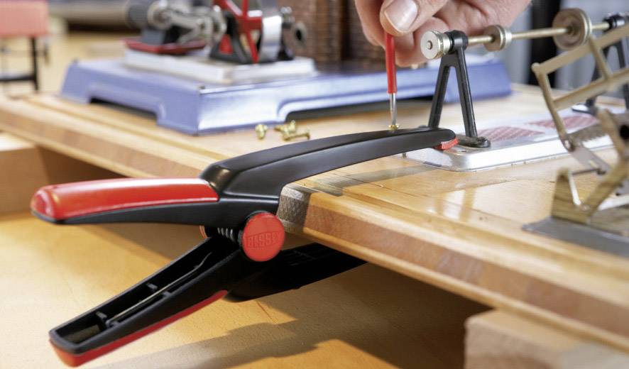 Osoba předvádí upínání modelářského materiálu červenou svěrkou a přesnými nástroji na pracovním stole.