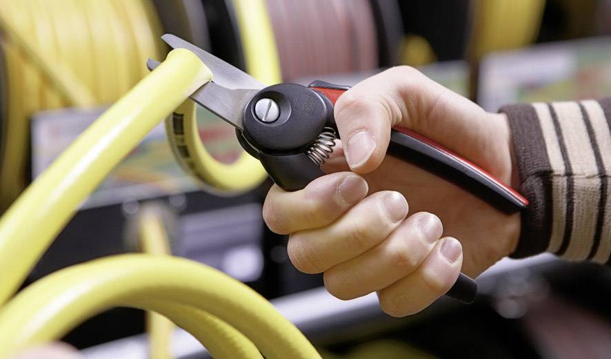 Osoba stříhá žlutou zahradní hadici nůžkami. V pozadí jsou viditelné další hadice a police.