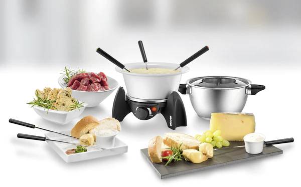 Stůl s fondue soupravou a příslušenstvím: sýrové fondue v hrnci, chléb, maso, omáčky a sýrová deska, ideální pro společné stolování.