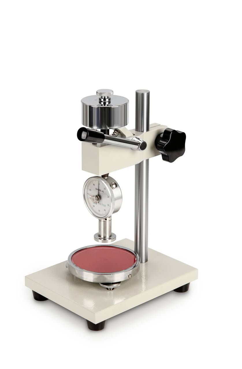 Zkušební přístroj na měření tvrdosti stojí na stole. Má kulatou měřicí plochu dole a ciferník pro zobrazení hodnot tvrdosti nahoře.