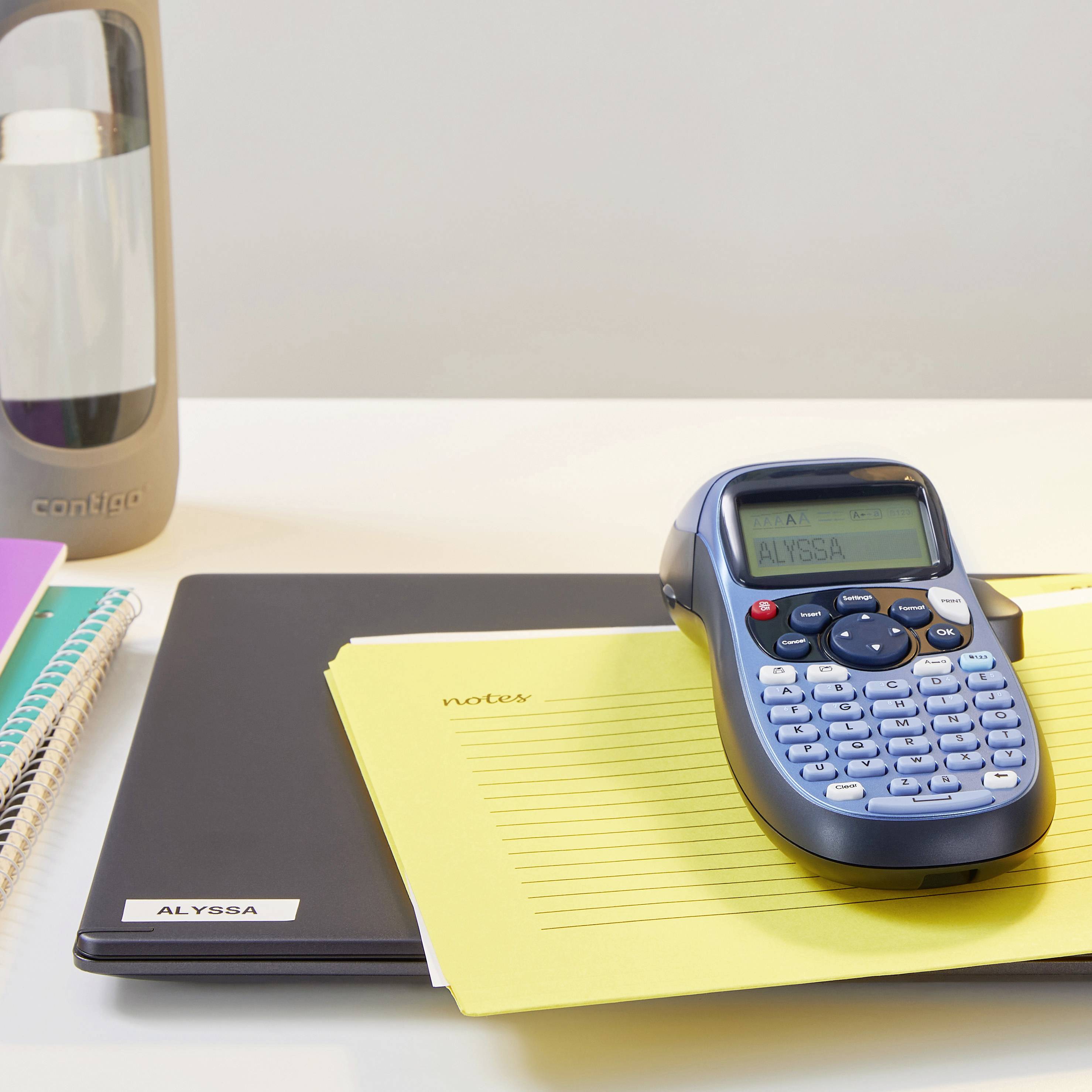 Poznámkový blok, žlutý zápisník s nápisem 'notes', tiskárna štítků a termohrnček na pracovním stole.