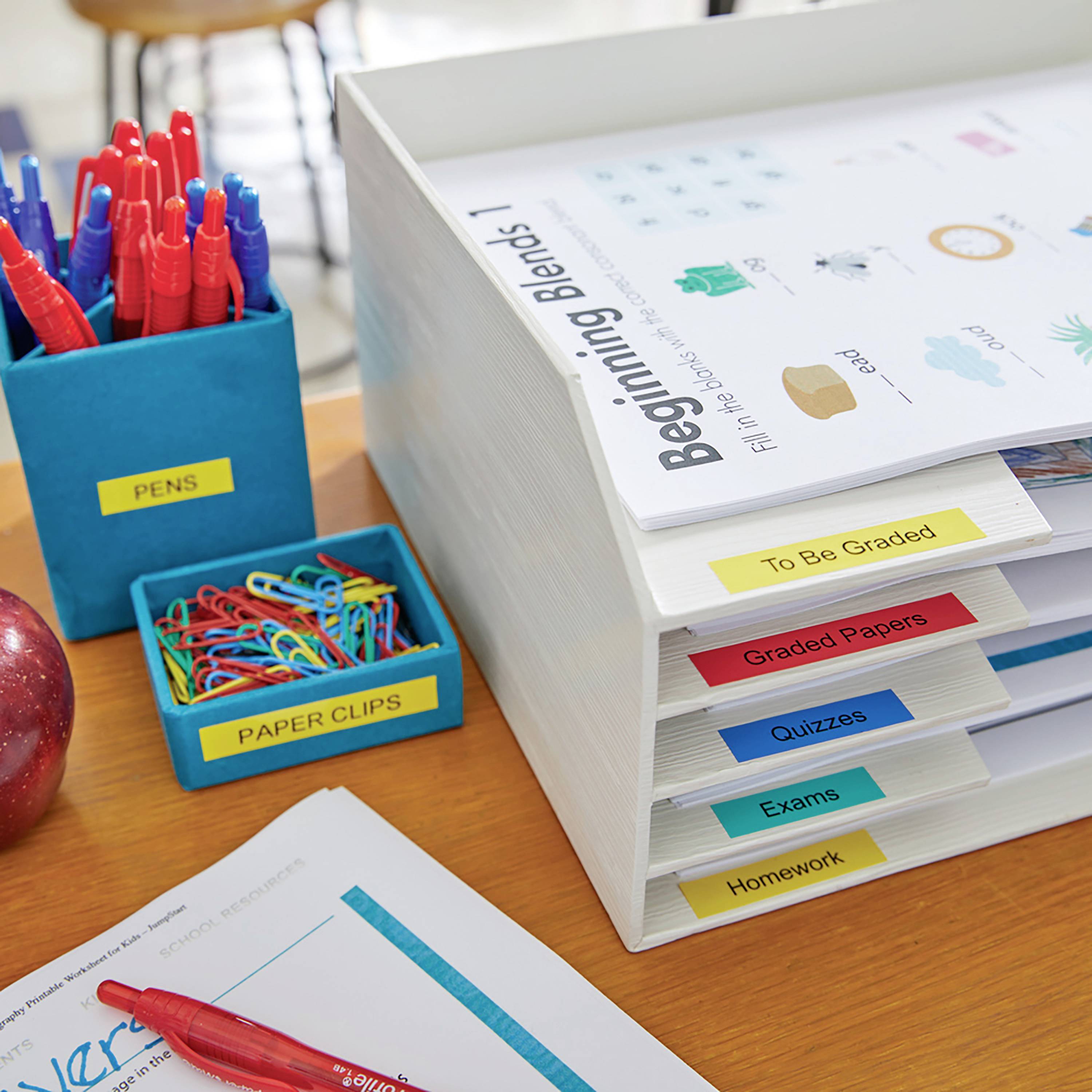 Kancelářský pracovní prostor s pořadači, které jsou označeny štítky 'Hodnocené práce', 'Zkoušky', 'Testy' a 'Domácí úkoly'. Tužky a kancelářské sponky v držácích.