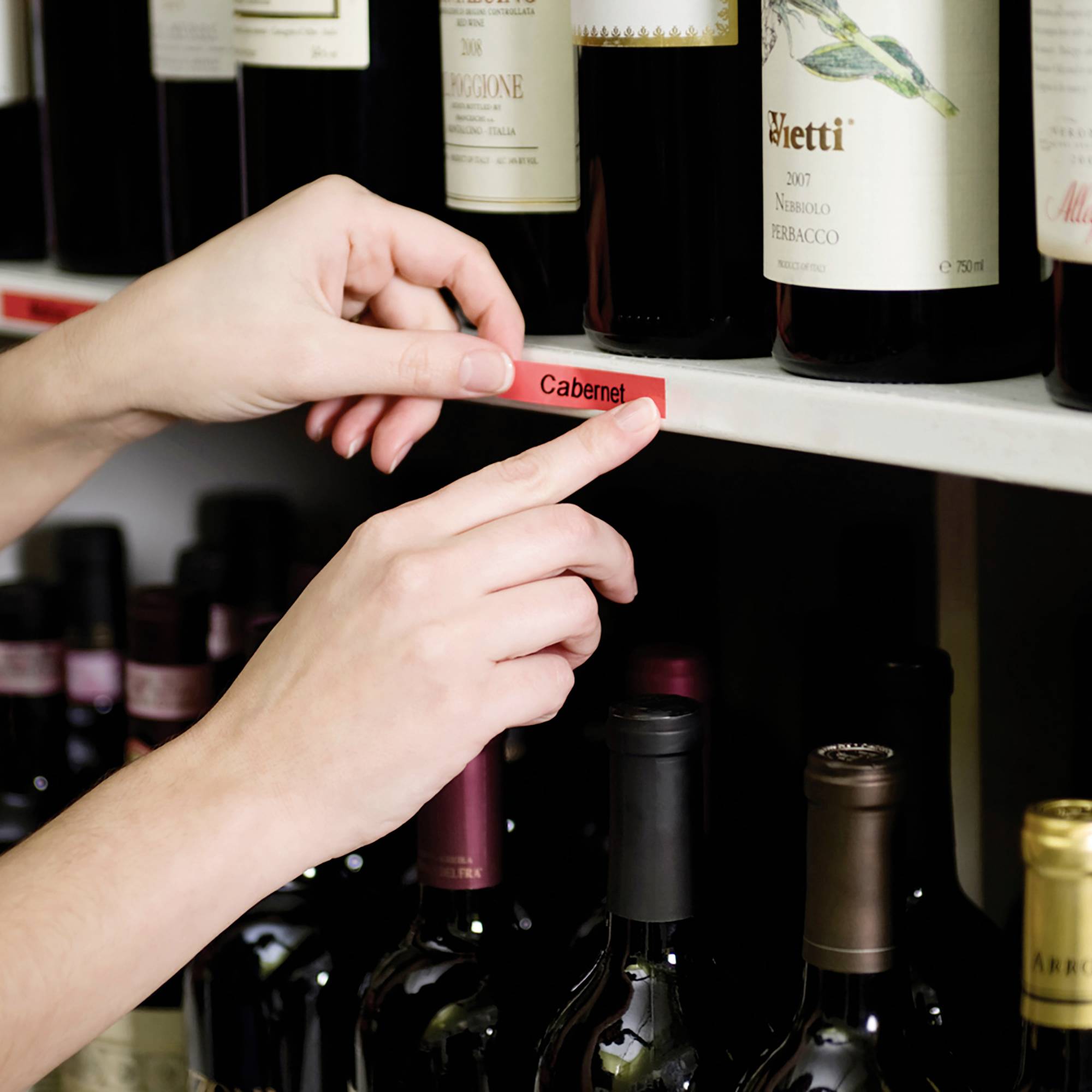 Ruka umisťuje červenou etiketu s nápisem 'Cabernet' na regál s vínem, aby označila láhve. V pozadí jsou viditelné láhve vína.