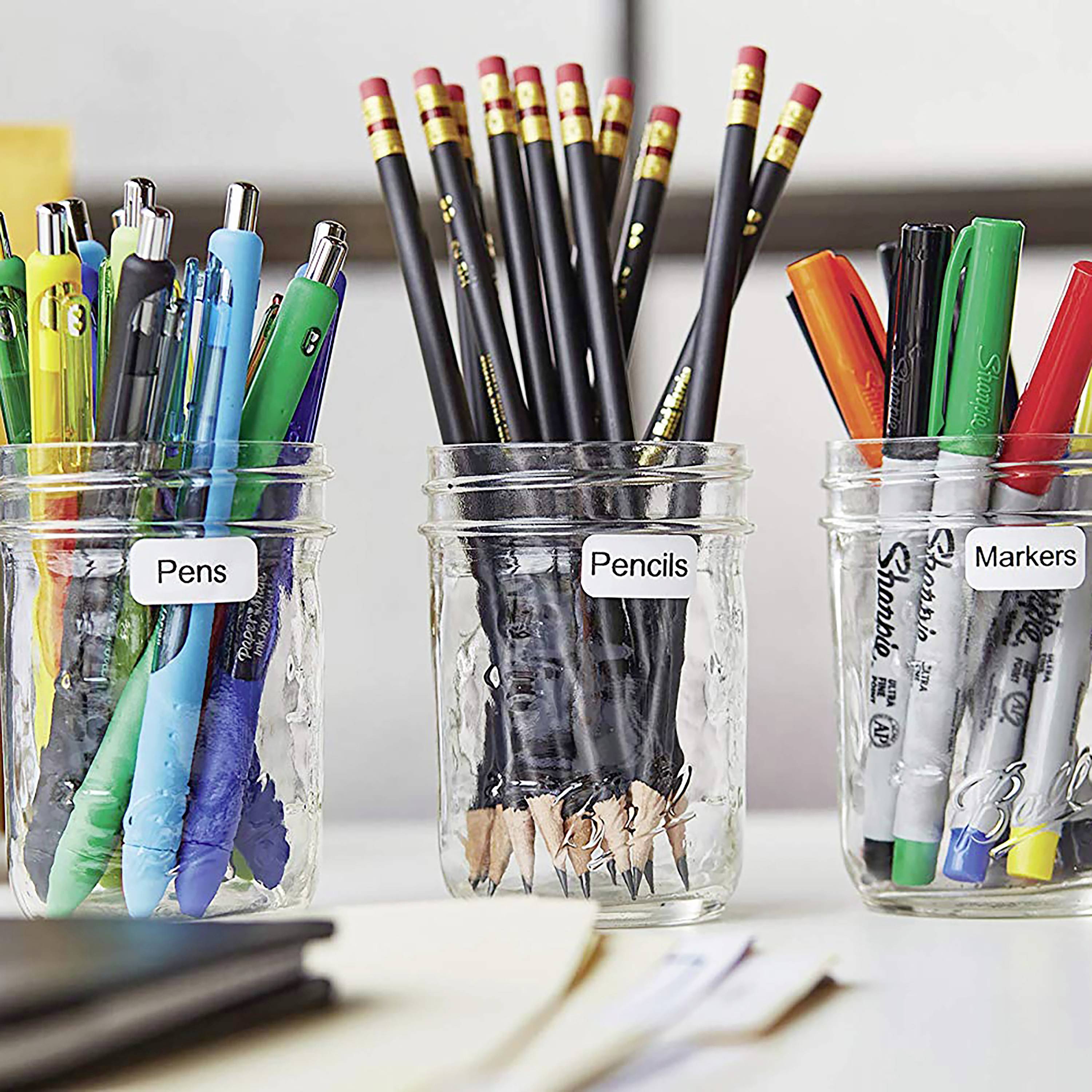 Tři skleněné nádoby s nápisy 'Pens', 'Pencils', 'Markers' stojí na stole, naplněné různými psacími potřebami.