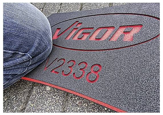 Koleno v modrých džínách je tisknuté proti červeno-šedé podložce, na které je reliéfně vyraženo slovo 'VIGOR' a 'V2338'.