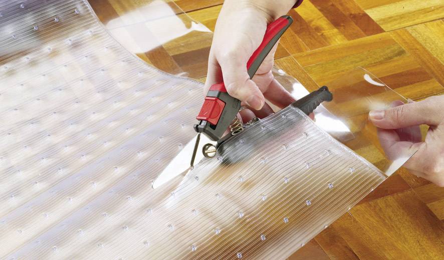 Osoba stříhá průhlednou plastovou fólii nůžkami na dřevěné podlaze.