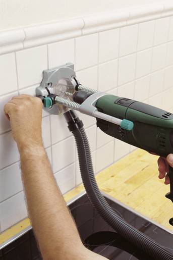 Ruka drží vrtačku s odsávacím zařízením a vrtá díru do bílé obkladové stěny. Žlutý pracovní povrch je v popředí.