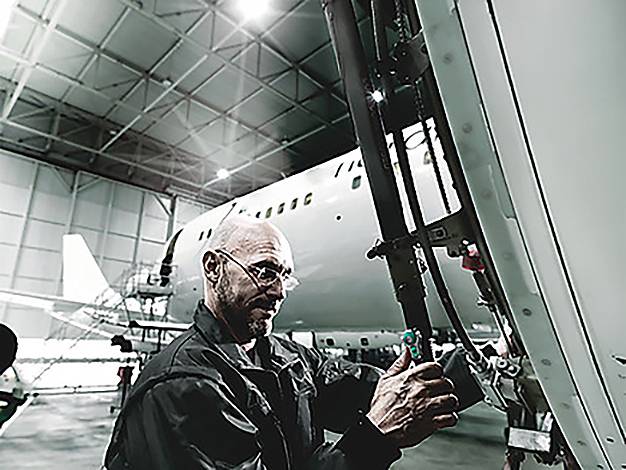 Mechanik pracuje na letadle uvnitř hangáru, s precizní soustředěností seřizuje vybavení, zatímco letadlo sedí v pozadí.