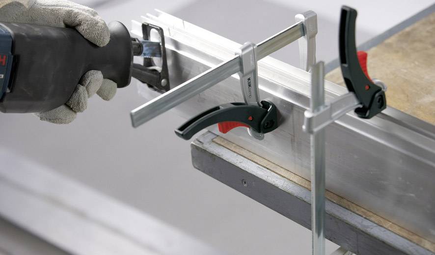 Ruka v rukavicích drží elektrickou pilu a řeže upevněný kovový díl, který je zajištěn svěrkami na stole.