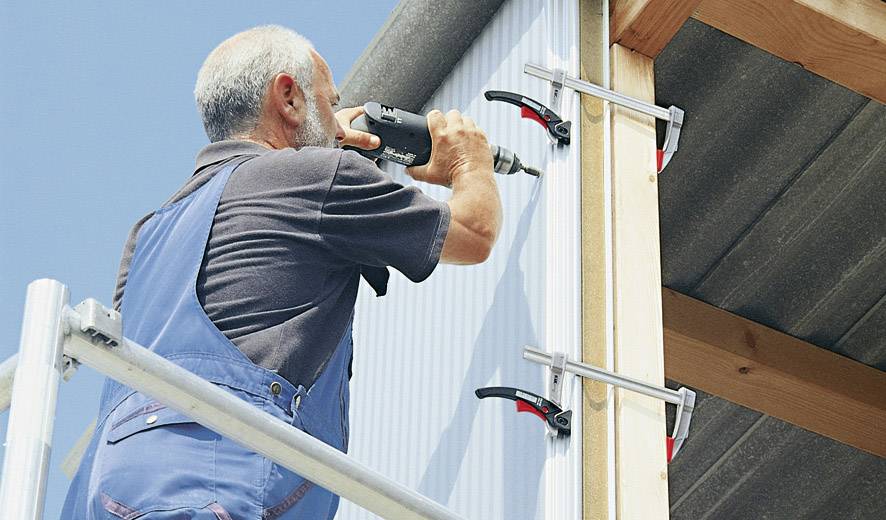Muž v pracovním oblečení upevňuje kovovou desku akumulátorovým šroubovákem na dřevěné konstrukci. Stojí na zdvižné plošině.