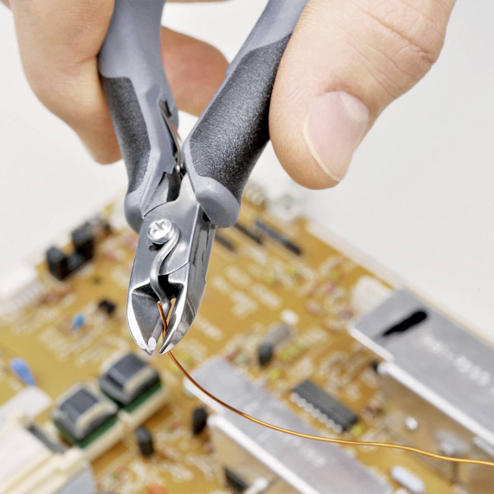 Jedna ruka drží štípací kleště, které řeže tenký drát nad elektronickou deskou osazenou různými komponenty.