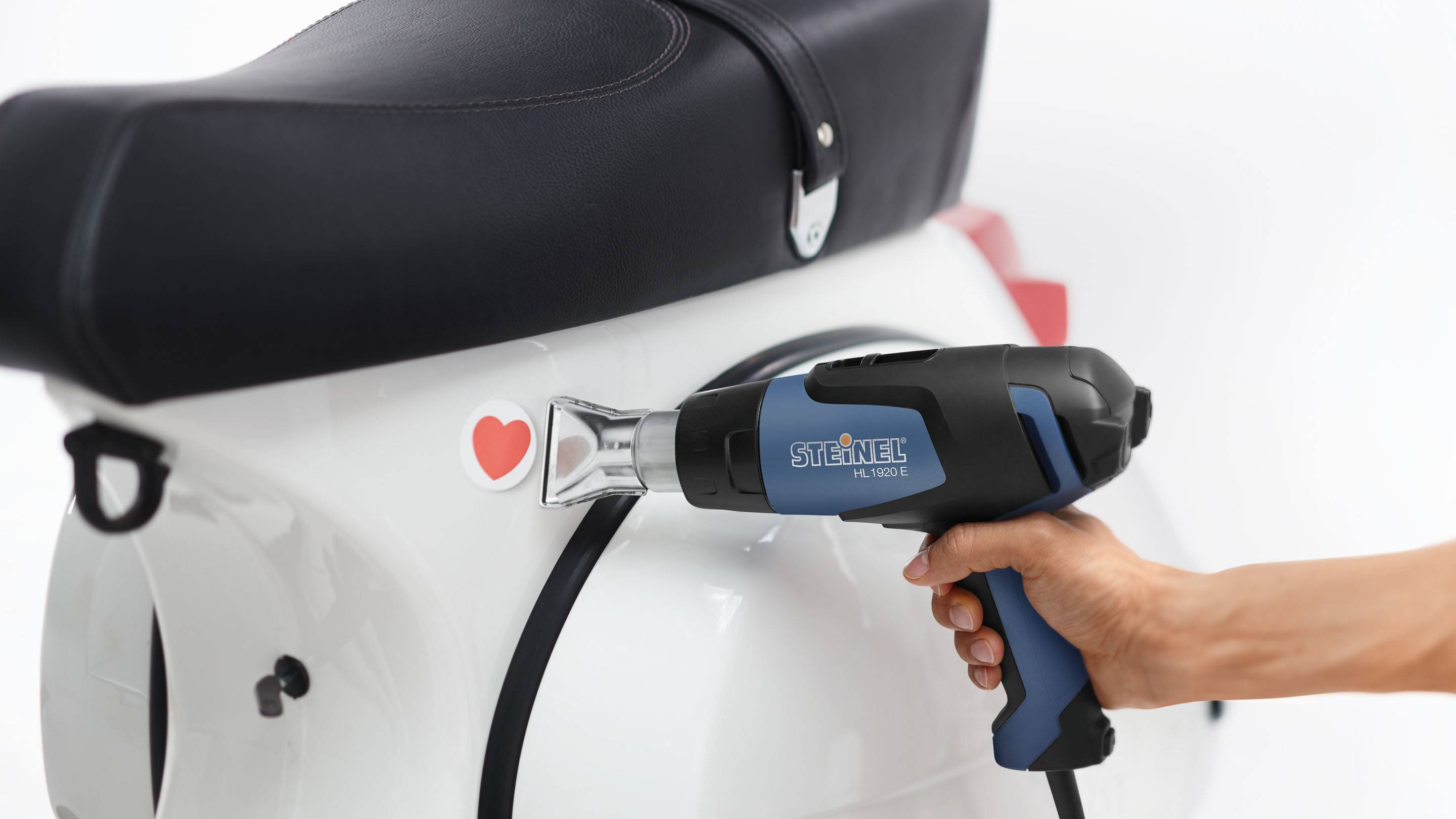 Osoba míří horkovzdušný ventilátor na bílou koloběžku s červenou nálepkou ve tvaru srdce.