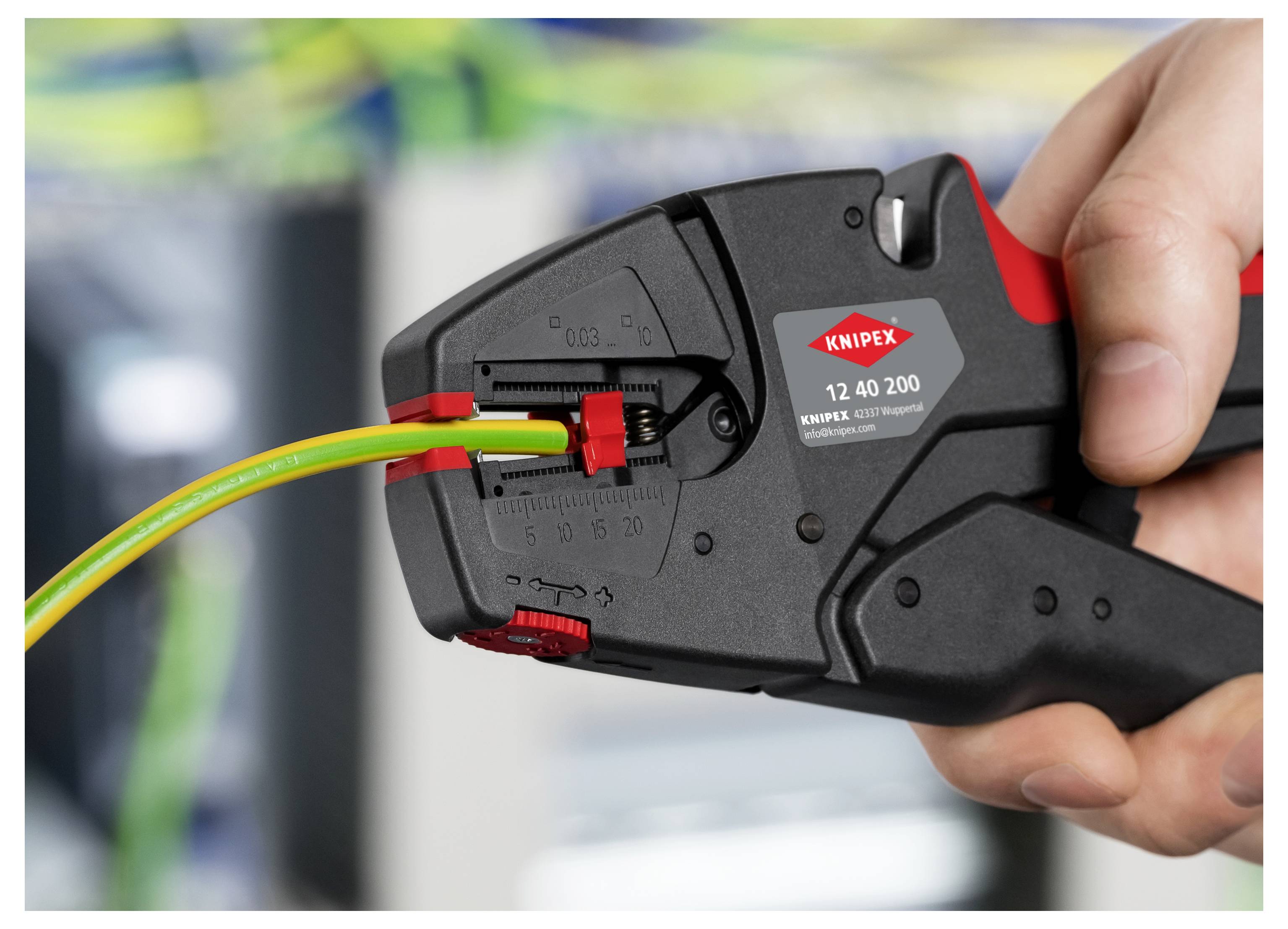 Ruka používající odizolovací kleště Knipex k odstranění izolace elektrického kabelu, obklopená rozmazanými kabely v pozadí.