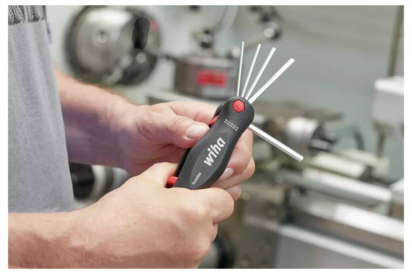 Osoba držící multitool s několika rozšířenými kovovými šestihranными klíči v nastavení dílny se strojním zařízením v pozadí.