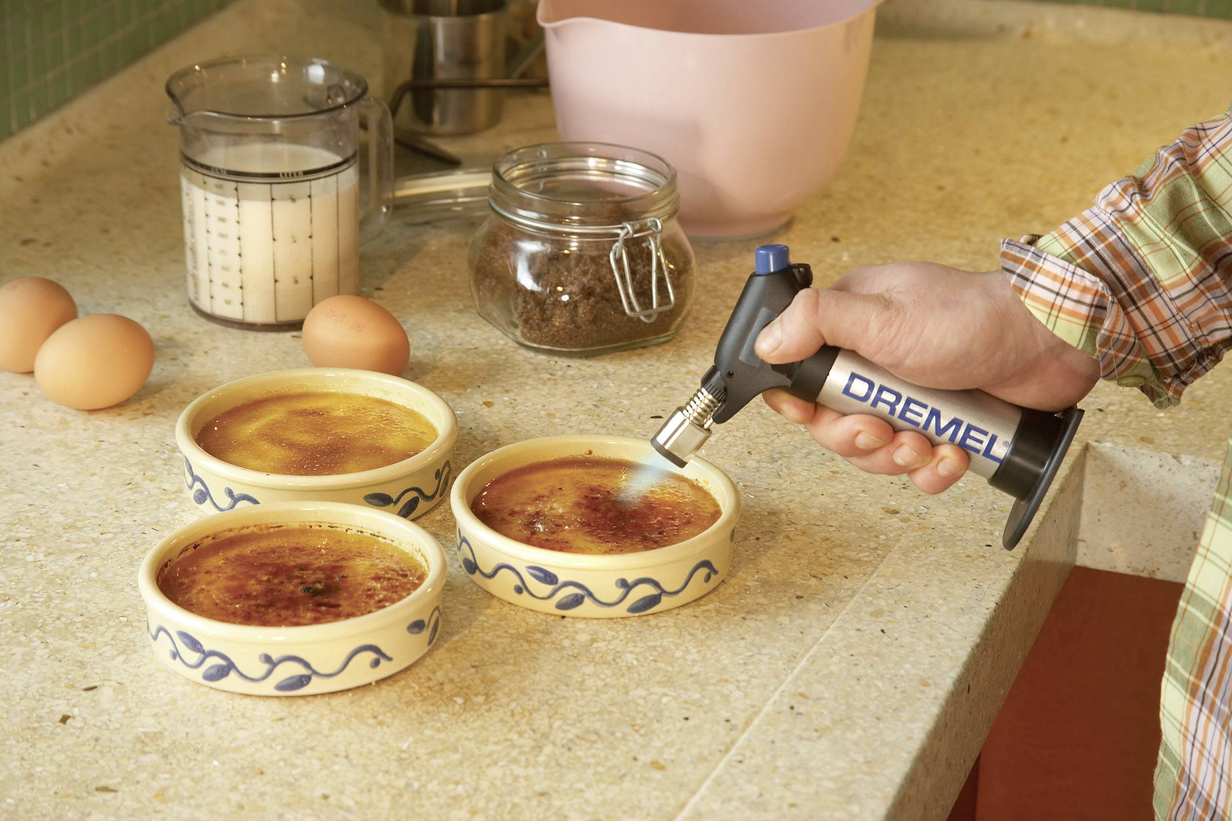 Osoba používá flambovací hořák, aby karamelizovala povrch tří misek s crème brûlée v kuchyni.