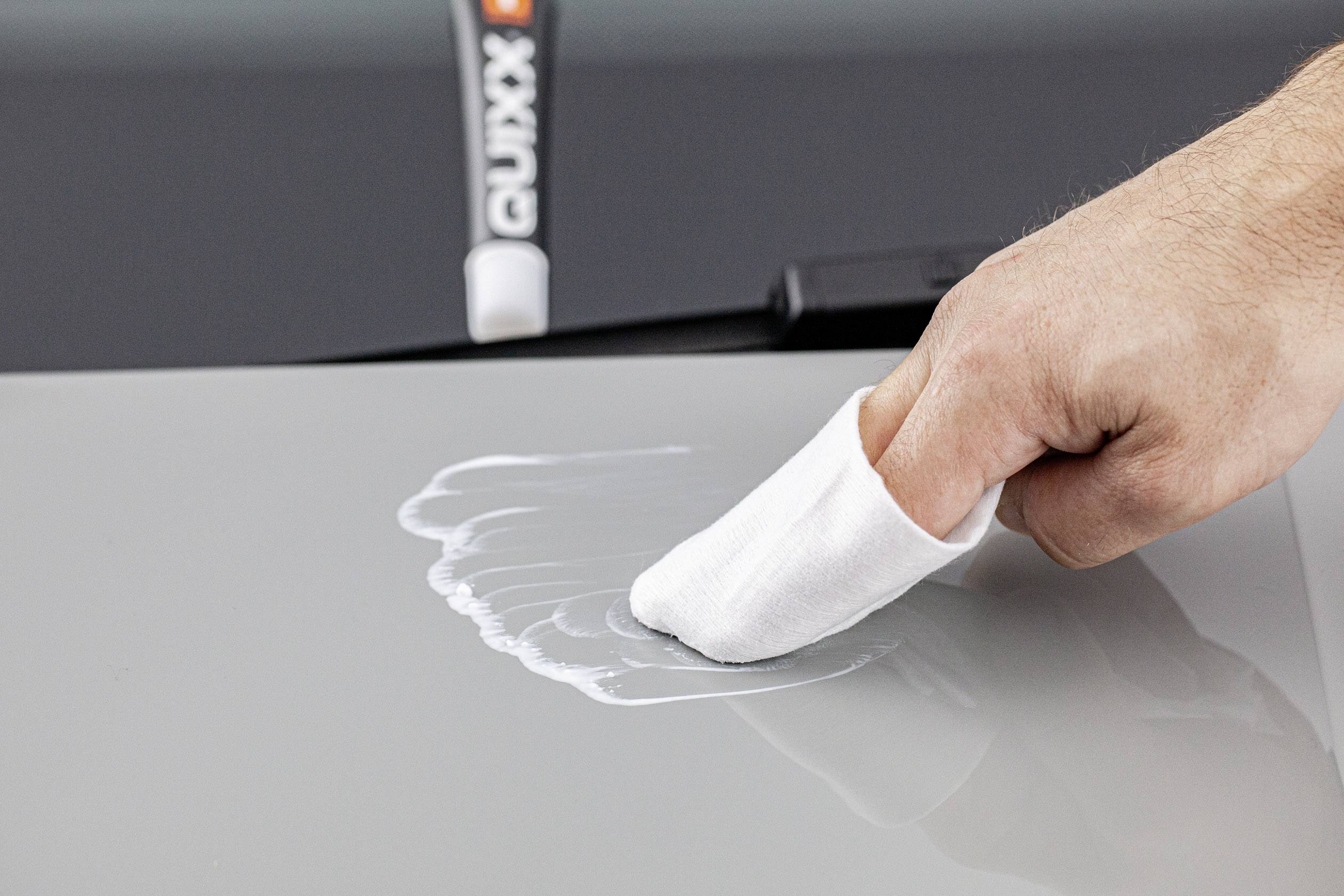 Ruka s bílou utěrkou leští bílou pastu na šedém autolaku. V pozadí stojí tuba s leštidlem.