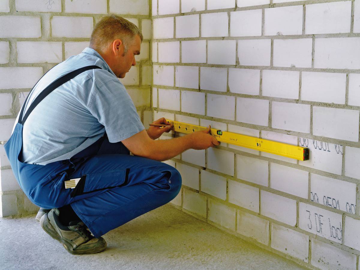 Řemeslník kontroluje vyrovnání cihlové zdi pomocí vodováhy, přičemž klečí na betonové podlaze.