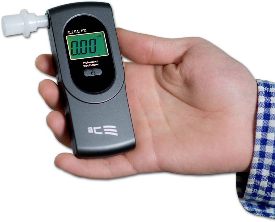 Jedna ruka drží přístroj na měření dechu alkoholu s digitálním displejem, který ukazuje '0,00'. Přístroj je připraven k použití a nevykazuje žádné hodnoty alkoholu.