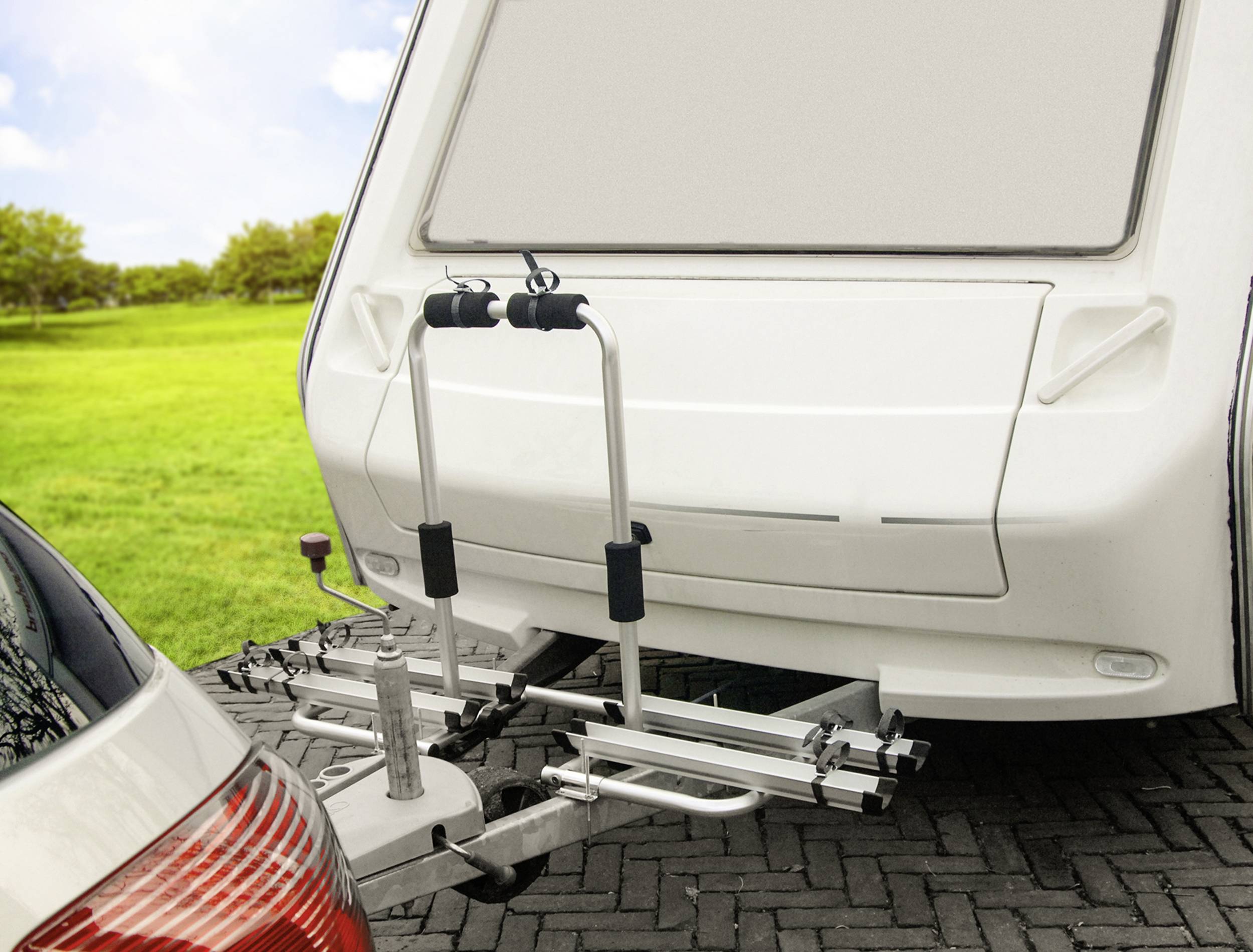 Auto s připojeným obytným přívěsem, na jehož zadní části je viditelný nosič jízdních kol. V pozadí je patrná zelená krajina.