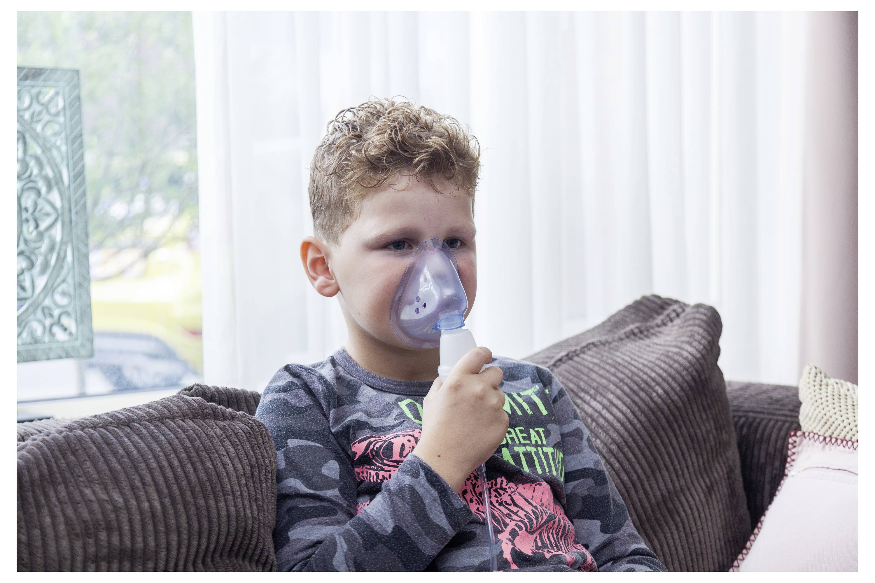 Malý chlapec sedí na pohovce a používá masku z nebulizéru pro léčbu dýchacích cest, což naznačuje zaměření na respirační zdraví nebo nemoc.